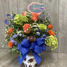 Football-themed bouquet with orange roses, green hydrangeas, and a blue bow in a sports container.