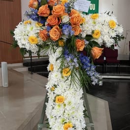 Large floral cross on an easel with white chrysanthemums, orange roses, and blue flowers