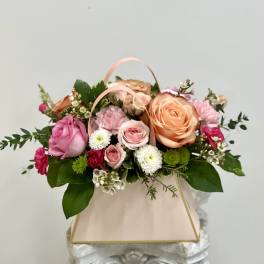 Pastel pink and peach flower arrangement with roses and mums in a blush gift bag-style container.