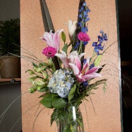 Tall mixed bouquet with pink lilies, blue flowers, and a hydrangea in a glass vase