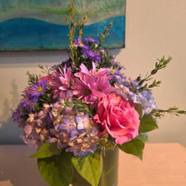 Compact arrangement of pink rose, purple daisies, and lavender hydrangeas in a clear glass cylinder vase