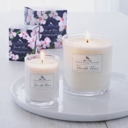 Two lit vanilla candles in glass jars with floral boxes behind them