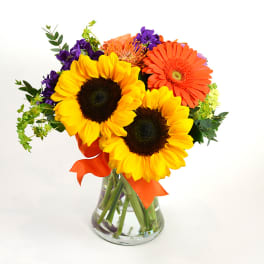 Bouquet of sunflowers, orange gerbera daisies, and purple flowers in a glass vase