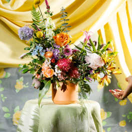 Colorful mixed flower arrangement in a terracotta pot on a draped table with yellow fabric backdrop.