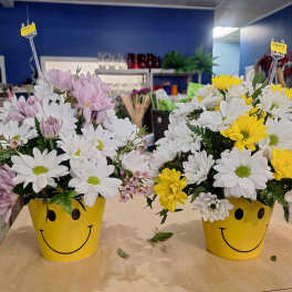Two daisy arrangements in yellow smiley-face pots, one with pink and white blooms and one with white and yellow blooms.