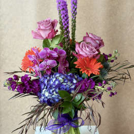 Mixed bouquet with pink roses, purple orchids, blue hydrangea, and orange gerbera daisies in a glass vase