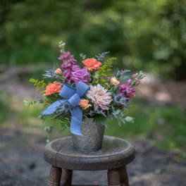 Mixed arrangement of peach carnations, lavender rose, and pink mum in a tin pot with a blue bow.