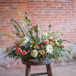 Low pastel arrangement of roses, mums, and daisies with an angel figurine in the center on a rustic stool