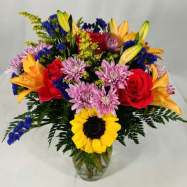 Bright mixed bouquet with sunflower, red roses, orange lilies, and purple mums in a clear glass vase