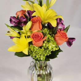 Bouquet of yellow lilies, orange roses, and purple flowers in a clear glass vase
