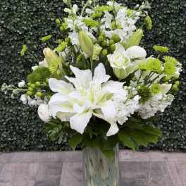 Tall white and green floral arrangement with lilies and mums in a patterned glass vase