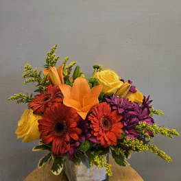 Colorful bouquet with orange lilies, red gerbera daisies, yellow roses, and purple flowers in a square vase