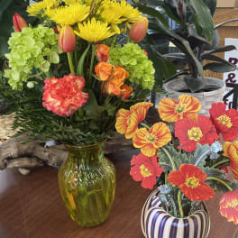 Bright mixed flower arrangement in a green glass vase beside a striped ceramic pot