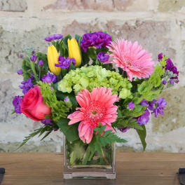 Pink gerbera daisies and roses in a clear glass vase with purple accents