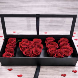 Red roses arranged in a black rectangular box with clear lid, surrounded by small red heart accents