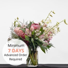 Pink protea and white orchids in a clear glass vase with text noting a 7-day advance order requirement.