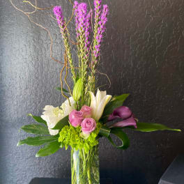 Tall vase arrangement with purple spikes, pink roses, white lilies, and dark purple calla lilies