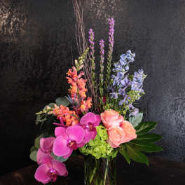 Tall modern arrangement with pink orchids, peach roses, blue delphinium and green hydrangea in a glass vase