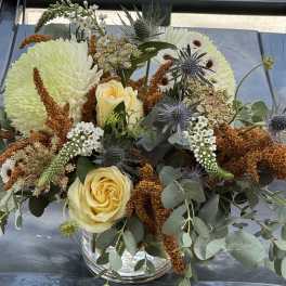 Bouquet of pale yellow roses, white chrysanthemums, and blue thistles in a glass vase