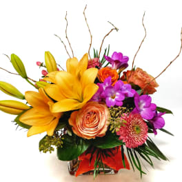 Colorful bouquet with yellow lilies, orange roses, and purple flowers in a glass vase