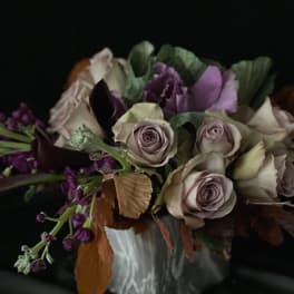 Moody arrangement of lavender roses, dark purple flowers, and kale in a white ceramic vase