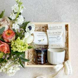 Floral gift box with roses and white blooms beside tea, caramels, fig spread, and a mug