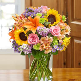 Mixed bouquet of sunflowers, roses, lilies, and purple flowers in a glass vase