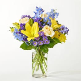Bouquet of yellow lilies, white roses, and blue-purple flowers in a clear glass vase