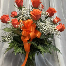 Orange roses in a glass vase with a large orange ribbon and white filler flowers.