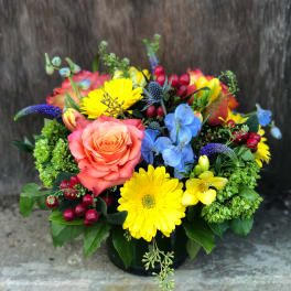 Colorful arrangement of yellow gerbera daisies, orange roses, blue delphinium and green blooms in a low glass vase