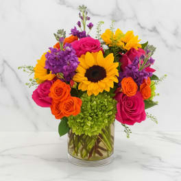 Bright mixed bouquet with roses, sunflowers, and purple blooms in a glass vase