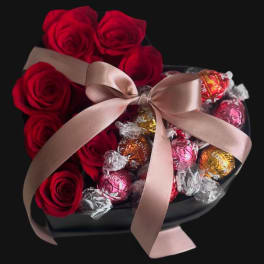 Red roses arranged with wrapped chocolates in a black box, tied with a pink ribbon.