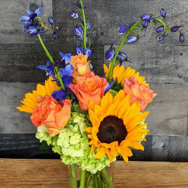 Tall glass vase with yellow sunflowers, orange roses, green hydrangeas, and blue delphinium.