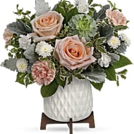Compact arrangement of peach roses, carnations, and white mums in a white ceramic pot on a wooden stand.