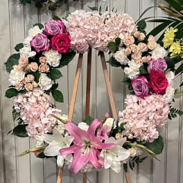 Heart-shaped floral wreath with pink hydrangeas, roses, and lilies on a wooden stand