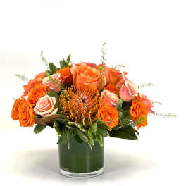 Low arrangement of orange and peach roses with a pincushion protea in a clear glass vase