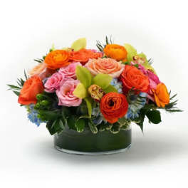 Low arrangement of orange and pink roses with green orchids in a clear glass cylinder vase