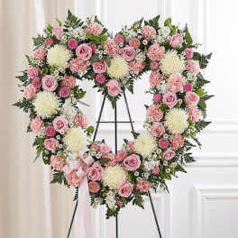 Heart-shaped standing wreath of pink roses, carnations, and white mums with a pink ribbon on an easel