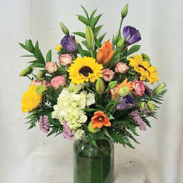Mixed bouquet of bright flowers including sunflowers, lilies, and roses in a clear glass vase.