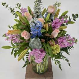 Tall pastel arrangement of pink roses, lilies, blue delphinium and stock in a clear glass vase