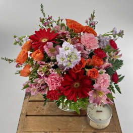 Bright mixed bouquet of red, pink, and orange flowers in a clear vase with a tin candle on a wooden crate
