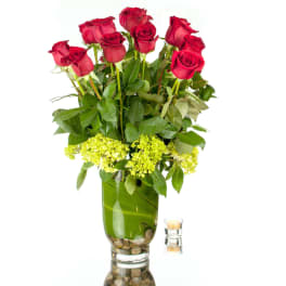 Tall arrangement of red roses with green blooms in a clear glass vase lined with a leaf and stones