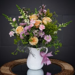 Peach roses and pink flowers in a white pitcher vase with a pink ribbon