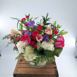 Mixed arrangement of pink roses, red carnations, purple blooms, and white hydrangeas in a glass vase