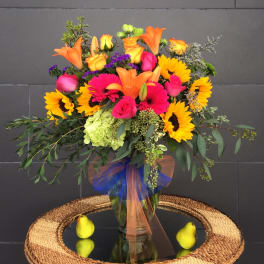 Bright mixed bouquet with sunflowers, roses, lilies, and gerbera daisies in a glass vase.