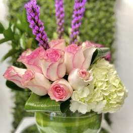 Low arrangement of pink roses, white hydrangea and tall purple spikes in a round glass bowl vase