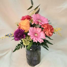 Colorful mixed arrangement of pink gerbera daisies, orange carnations, and yellow rose in a tinted glass jar