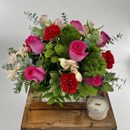 Arrangement of hot pink roses, red carnations, and green accents in a white box with a small candle beside it.