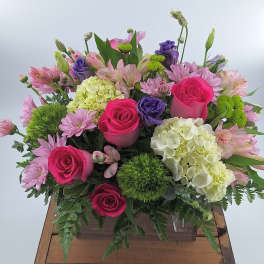 Mixed arrangement of hot pink roses, white hydrangeas, and purple blooms in a clear rectangular vase