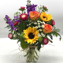 Mixed bouquet with sunflowers, orange roses, purple flowers, and white daisies in a glass vase